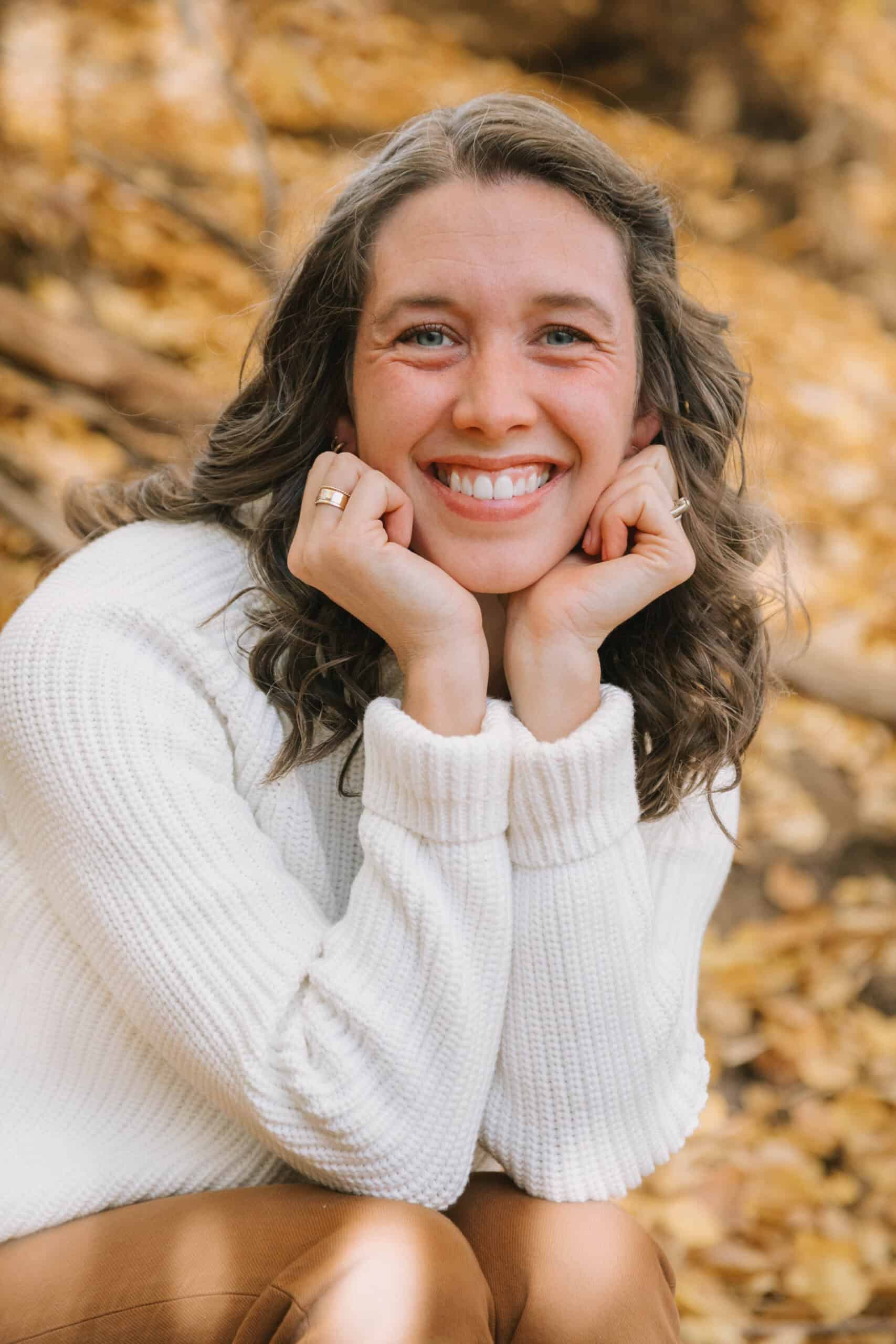 A photo of Libby Bloom with her hands framing her face as she smiles at the camera. She's wearing a white sweater and there are yellow leaves in the background.