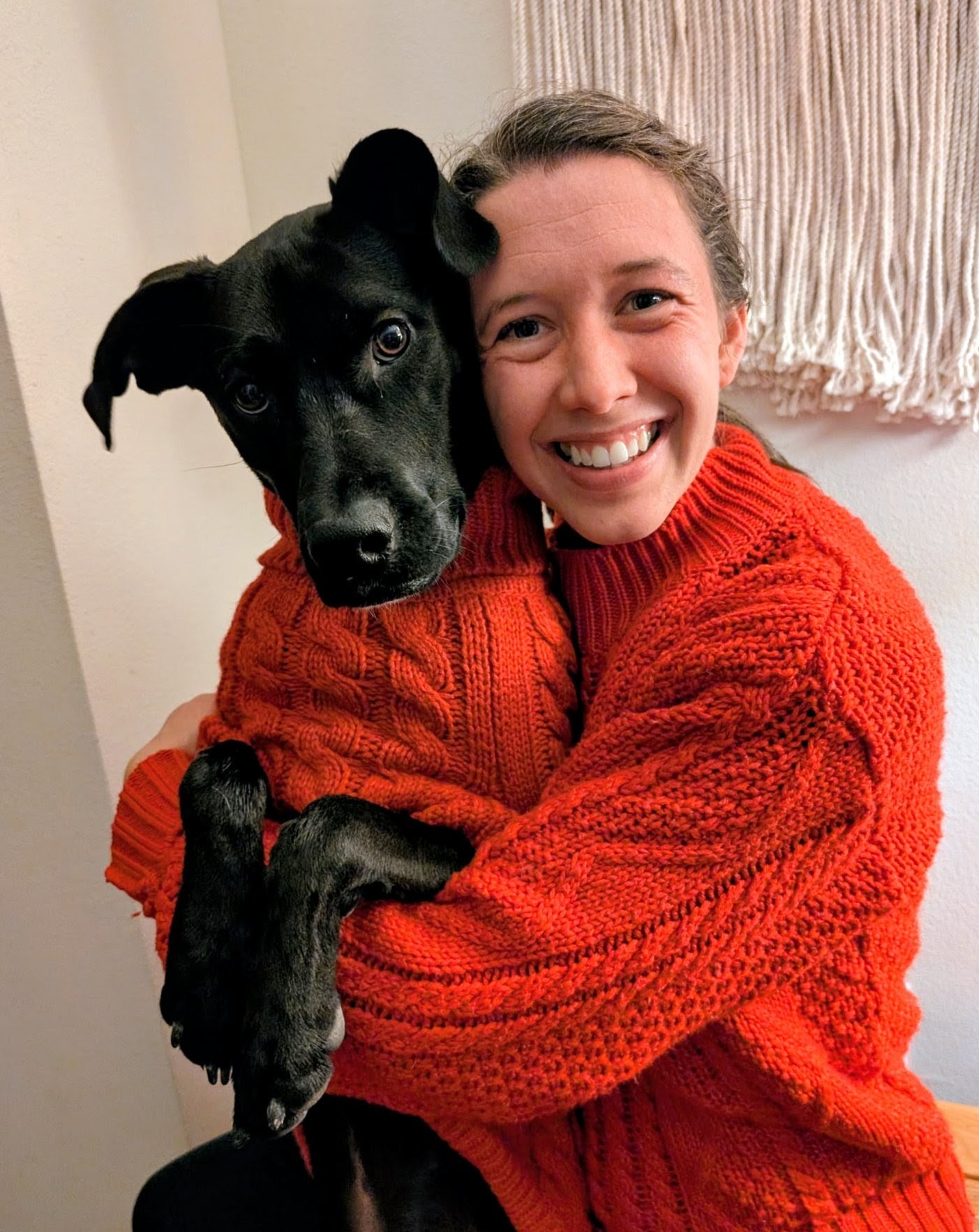 A girl wearing a red sweater smiling with a black dog wearing a red sweater
