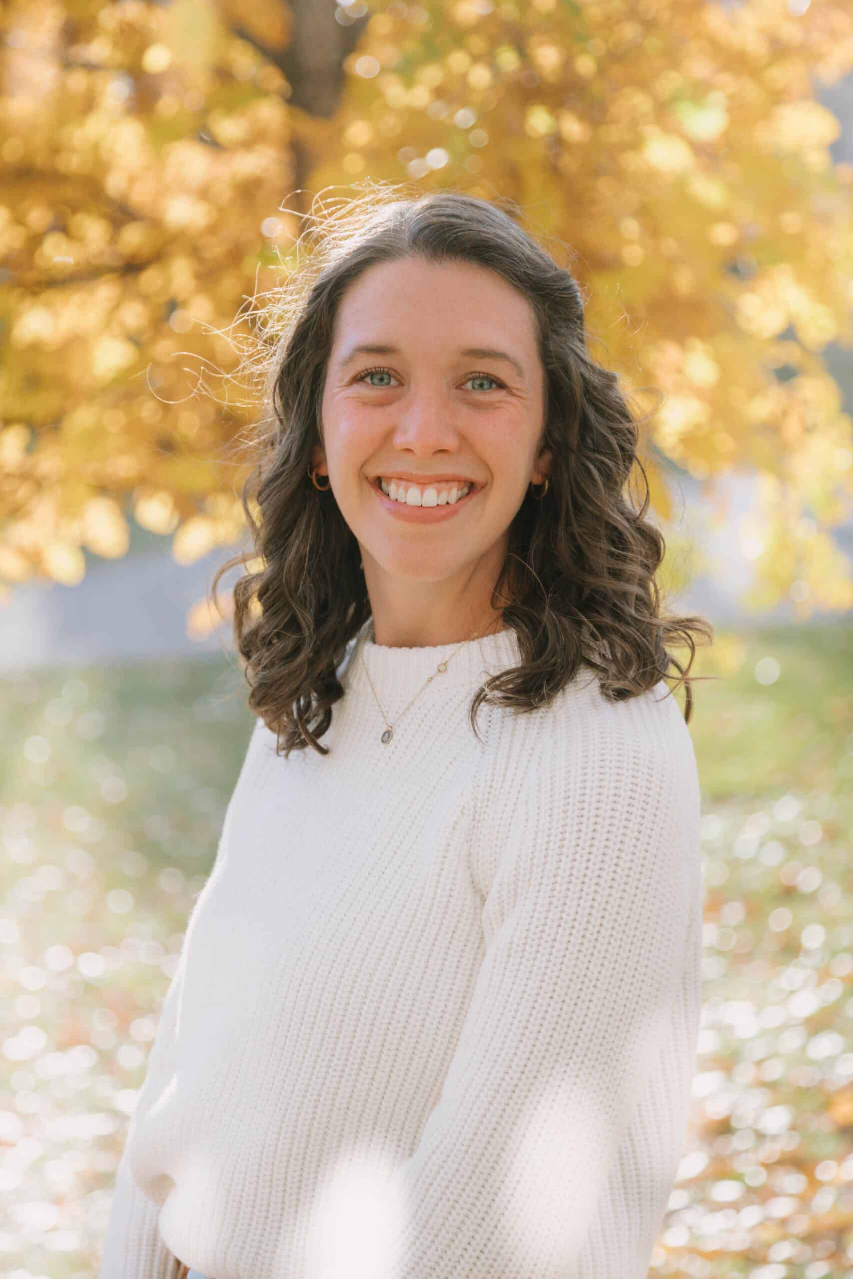 A photo of Libby Bloom looking at the camera wearing a white sweater with yellow leaves in the background
