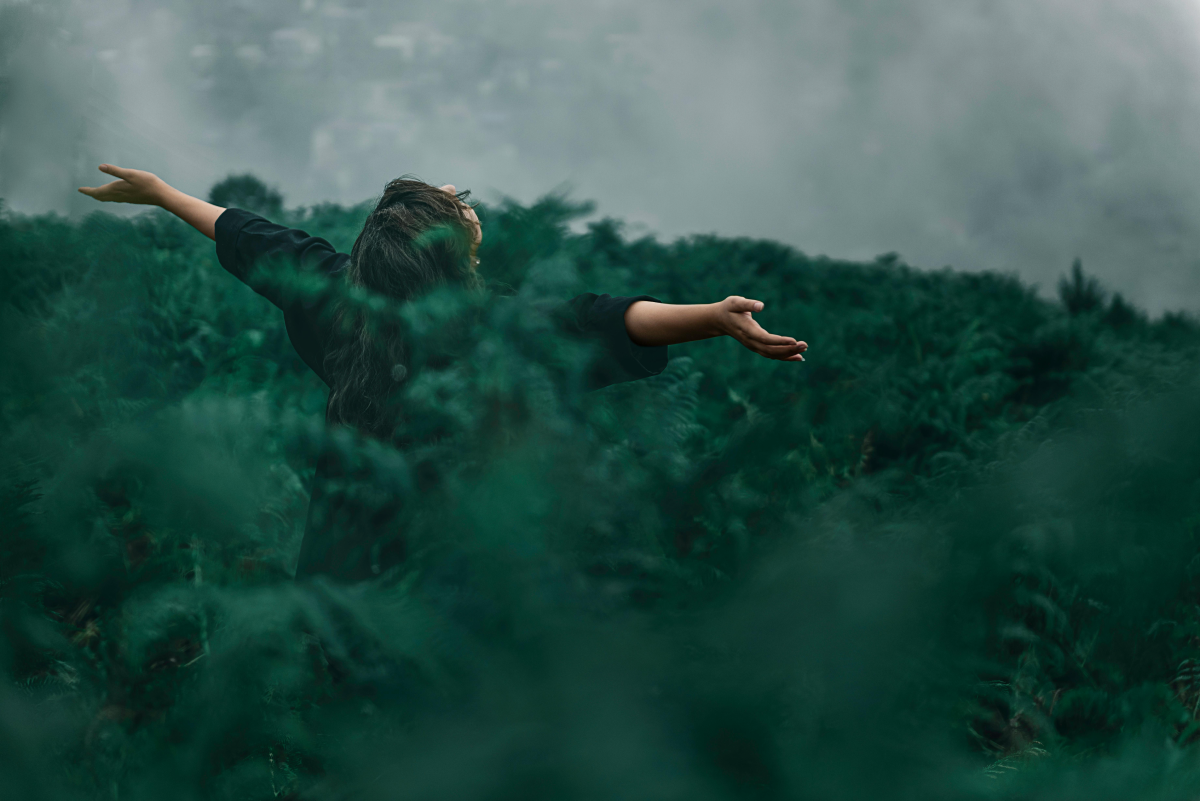 woman with her arms open to the sky. She's facing away from the camera and is standing in a field of ferns